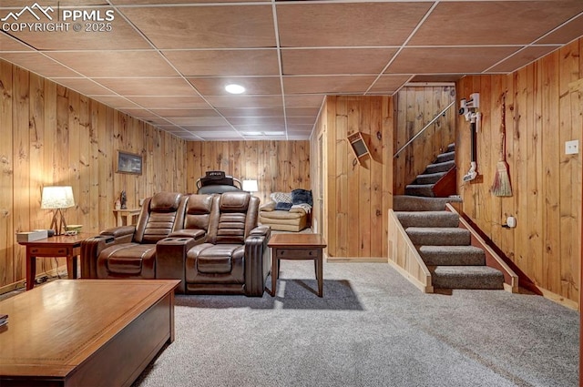 carpeted living area with a paneled ceiling, wood walls, stairway, and baseboards