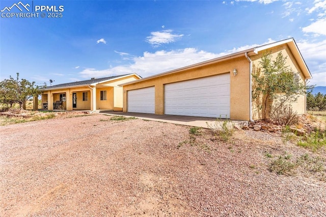 ranch-style house featuring dirt driveway, covered porch, a garage, and stucco siding