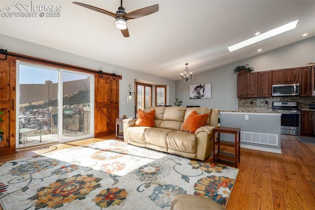 living area with a barn door, visible vents, lofted ceiling with skylight, wood finished floors, and ceiling fan with notable chandelier