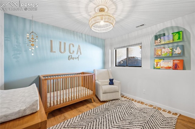 bedroom with a crib, baseboards, visible vents, wood finished floors, and a notable chandelier