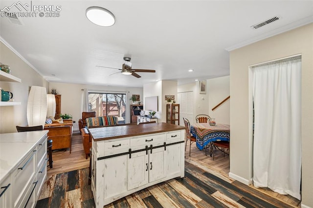 kitchen with dark wood-type flooring, white cabinetry, a ceiling fan, visible vents, and crown molding