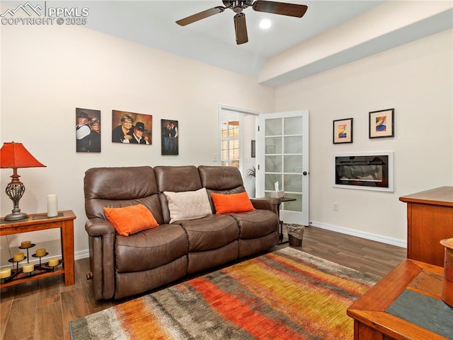 living area featuring a glass covered fireplace, wood finished floors, a ceiling fan, and baseboards