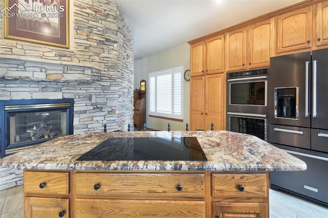 kitchen featuring light stone countertops, double wall oven, black electric cooktop, a stone fireplace, and stainless steel refrigerator with ice dispenser