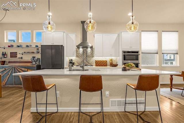 kitchen featuring double oven, a kitchen breakfast bar, wall chimney range hood, freestanding refrigerator, and light wood finished floors