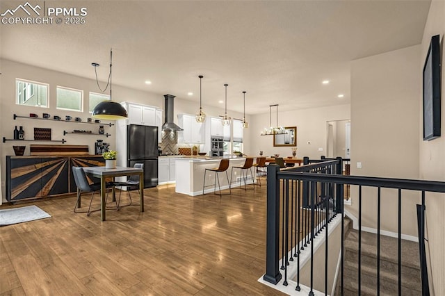 kitchen featuring wood finished floors, freestanding refrigerator, wall chimney range hood, a kitchen bar, and white cabinetry