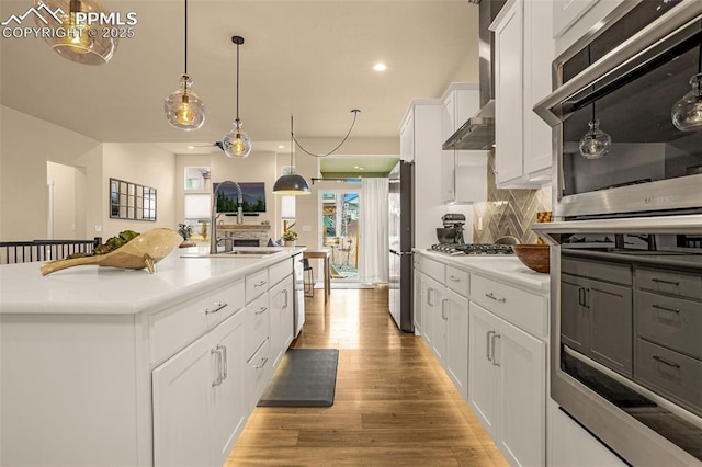 kitchen featuring light wood finished floors, tasteful backsplash, a center island with sink, wall chimney exhaust hood, and appliances with stainless steel finishes