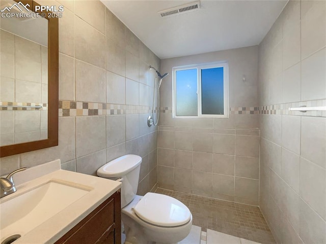 bathroom featuring toilet, vanity, visible vents, tile walls, and tiled shower