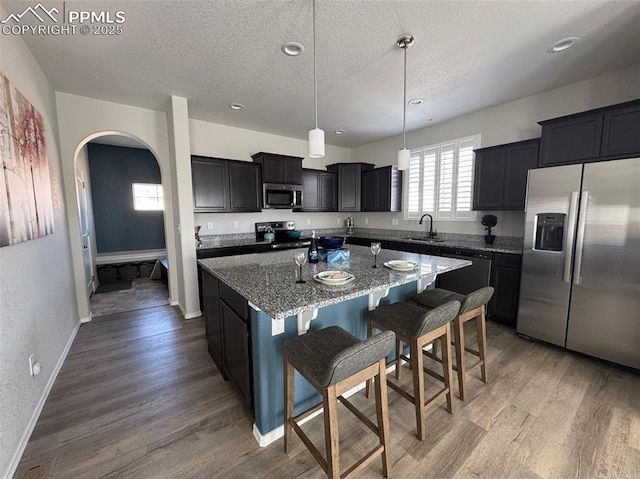 kitchen with arched walkways, a center island with sink, wood finished floors, appliances with stainless steel finishes, and light stone countertops