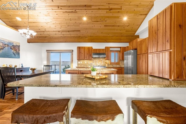 kitchen featuring brown cabinetry, wooden ceiling, a breakfast bar, freestanding refrigerator, and under cabinet range hood