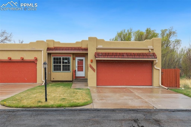 pueblo-style house with an attached garage, fence, concrete driveway, stucco siding, and a front lawn