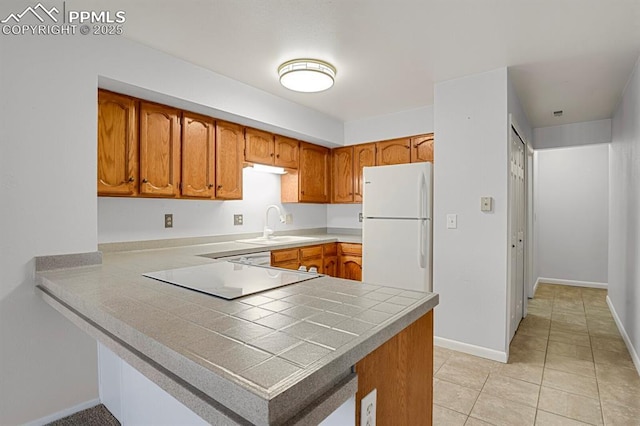 kitchen with brown cabinetry, freestanding refrigerator, light tile patterned flooring, a sink, and a peninsula