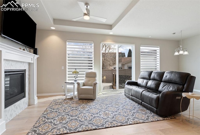 living area featuring ceiling fan with notable chandelier, a high end fireplace, baseboards, light wood-type flooring, and a tray ceiling