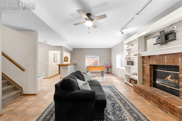 living room featuring rail lighting, stairway, a ceiling fan, a brick fireplace, and light tile patterned flooring