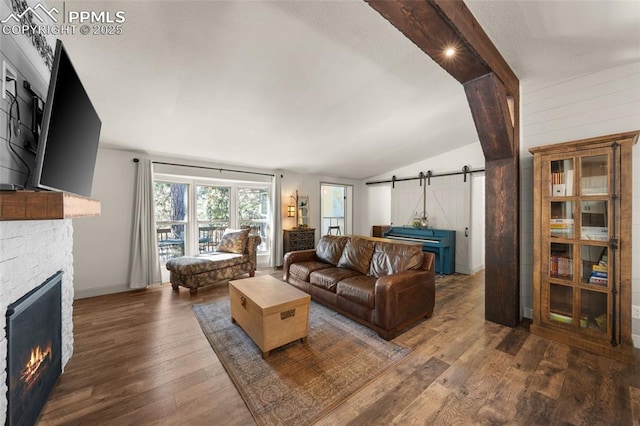 living room featuring wood finished floors, a barn door, a stone fireplace, baseboards, and vaulted ceiling with beams
