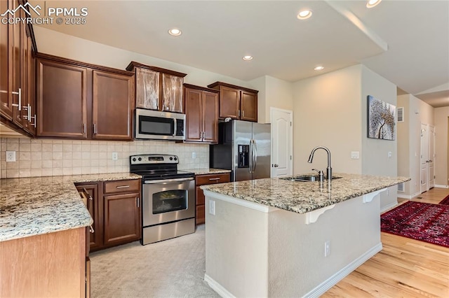 kitchen featuring a center island with sink, backsplash, appliances with stainless steel finishes, a sink, and light stone countertops