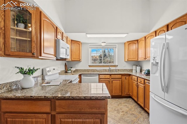 kitchen with white appliances, a sink, light stone countertops, brown cabinetry, and glass insert cabinets