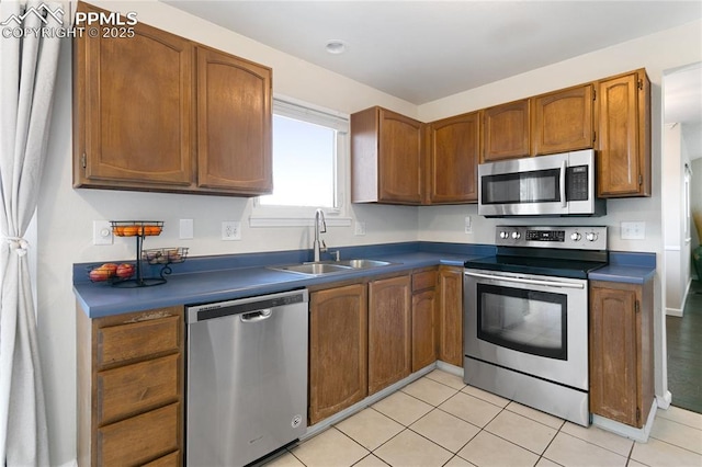 kitchen with brown cabinets, stainless steel appliances, dark countertops, light tile patterned flooring, and a sink