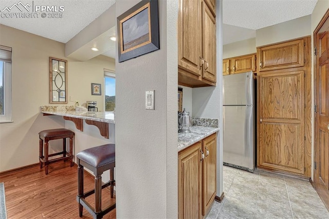 kitchen featuring baseboards, freestanding refrigerator, light stone countertops, a textured ceiling, and a kitchen bar