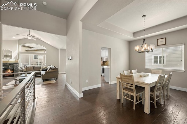 dining space featuring ceiling fan with notable chandelier, a fireplace, baseboards, dark wood-style floors, and a raised ceiling