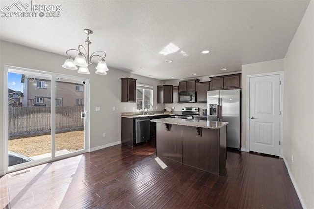 kitchen with dark brown cabinetry, stainless steel appliances, a kitchen island, light stone countertops, and dark wood finished floors