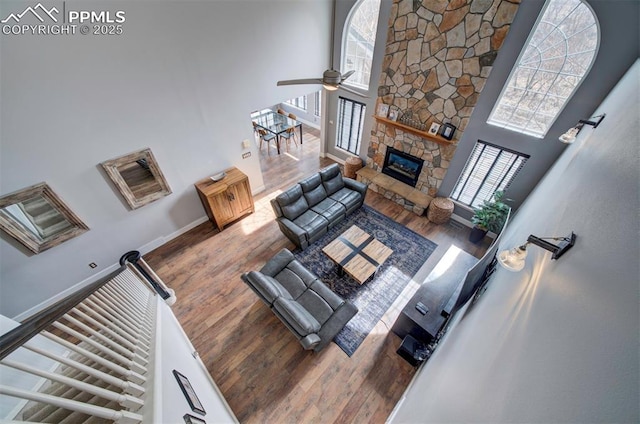 living area with a high ceiling, a ceiling fan, a stone fireplace, wood finished floors, and baseboards