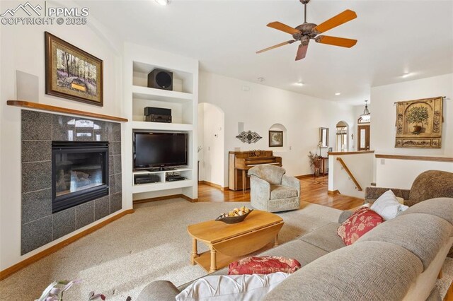 living area featuring baseboards, arched walkways, a tile fireplace, ceiling fan, and built in shelves