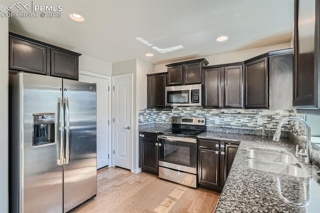 kitchen featuring appliances with stainless steel finishes, light wood-style floors, a sink, dark brown cabinetry, and dark stone counters