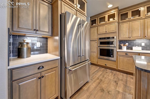 kitchen featuring stainless steel appliances, glass insert cabinets, backsplash, and light wood-style flooring