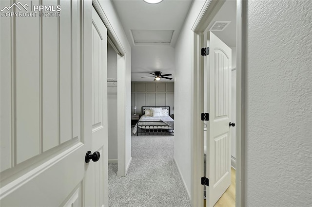 corridor featuring a textured wall, light colored carpet, visible vents, baseboards, and attic access