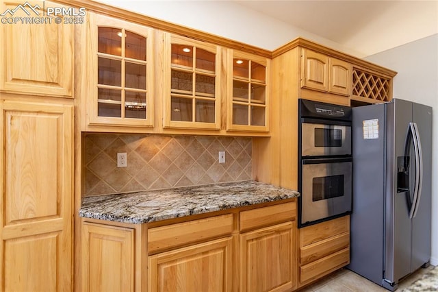 kitchen featuring light stone counters, light brown cabinetry, stainless steel appliances, glass insert cabinets, and tasteful backsplash