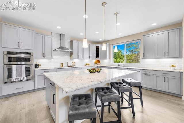 kitchen with wall chimney exhaust hood, light wood-style flooring, appliances with stainless steel finishes, gray cabinetry, and a kitchen bar