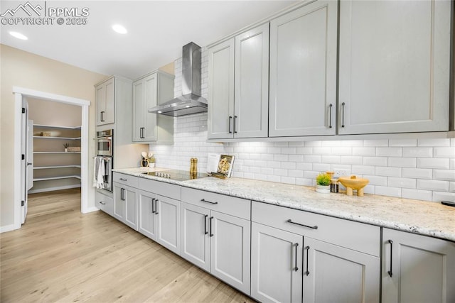 kitchen with black electric stovetop, light stone counters, wall chimney range hood, backsplash, and light wood finished floors