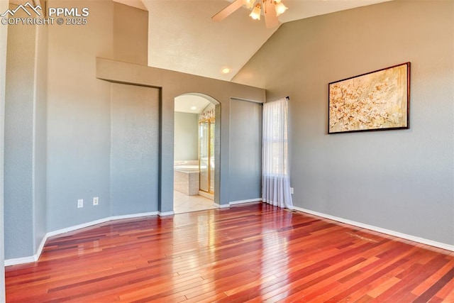 empty room featuring arched walkways, hardwood / wood-style floors, ceiling fan, high vaulted ceiling, and baseboards