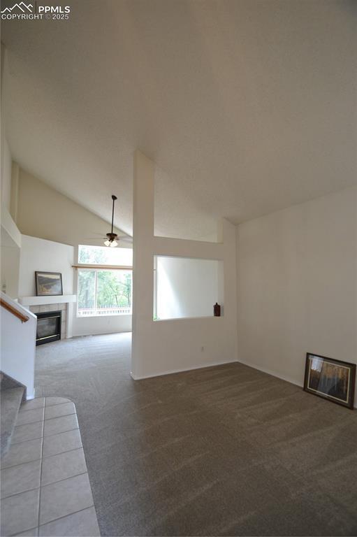 unfurnished living room featuring carpet, a tiled fireplace, ceiling fan, vaulted ceiling, and stairs