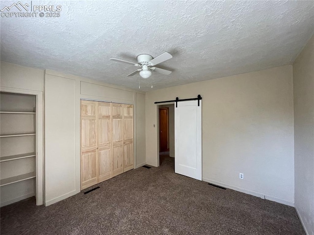 unfurnished bedroom featuring two closets, dark carpet, a barn door, ceiling fan, and a textured ceiling