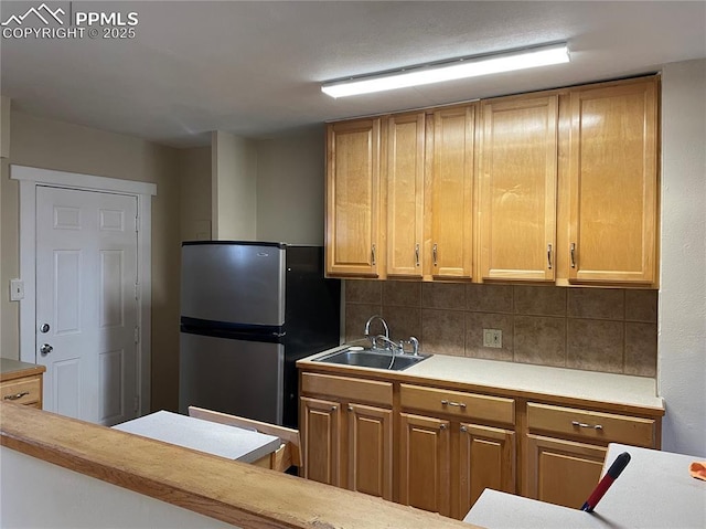 kitchen featuring a sink, light countertops, backsplash, freestanding refrigerator, and brown cabinetry