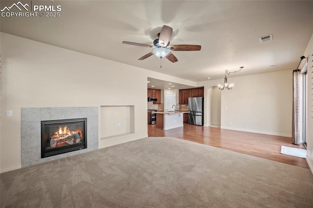 unfurnished living room featuring visible vents, baseboards, a tiled fireplace, light colored carpet, and ceiling fan with notable chandelier