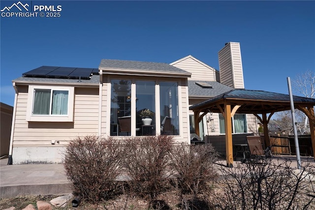 rear view of house featuring a shingled roof, a chimney, a gazebo, a patio area, and roof mounted solar panels