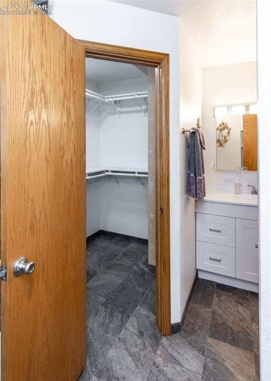 bathroom featuring stone finish floor, vanity, a walk in closet, and baseboards