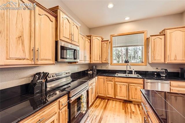 kitchen with light brown cabinets, a sink, dark countertops, stainless steel appliances, and light wood-style floors