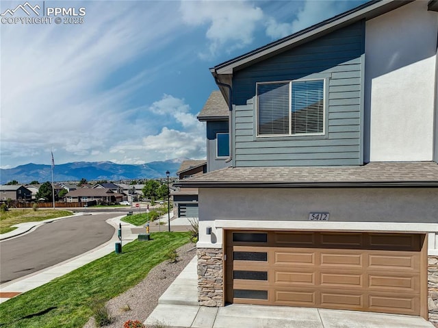 view of side of home with driveway, stone siding, a mountain view, an attached garage, and a shingled roof