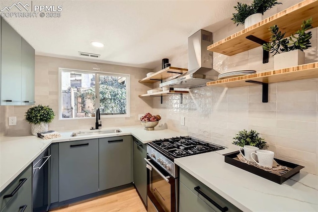 kitchen featuring open shelves, wall chimney range hood, stainless steel range with gas stovetop, gray cabinets, and a sink