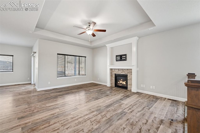 unfurnished living room featuring a stone fireplace, a tray ceiling, wood finished floors, and baseboards