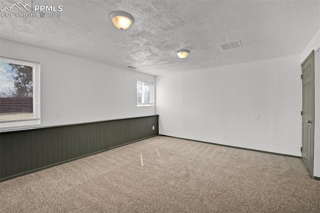 empty room featuring a textured ceiling, carpet, visible vents, and wainscoting