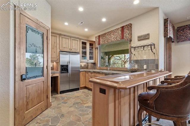 kitchen featuring tasteful backsplash, stainless steel refrigerator with ice dispenser, a peninsula, stone finish flooring, and glass insert cabinets