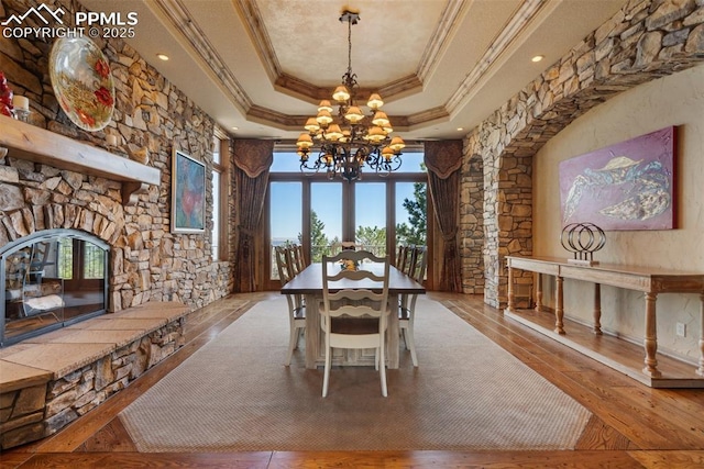 dining space with crown molding, hardwood / wood-style flooring, a raised ceiling, a chandelier, and a stone fireplace