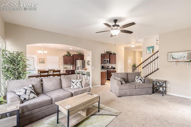 living room featuring stairs, light carpet, baseboards, arched walkways, and ceiling fan with notable chandelier
