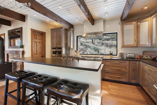 kitchen featuring appliances with stainless steel finishes, wall chimney range hood, dark countertops, and beamed ceiling