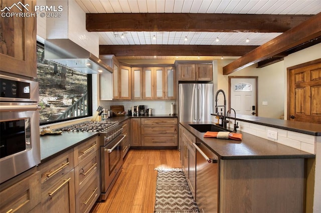 kitchen featuring dark countertops, appliances with stainless steel finishes, a sink, light wood-style floors, and wall chimney exhaust hood