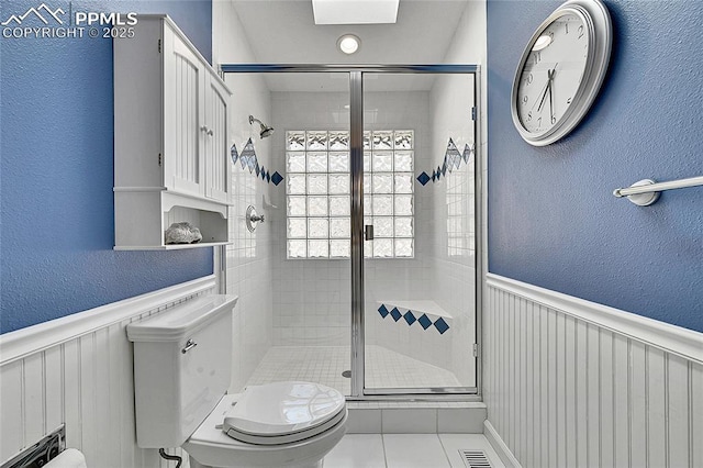 bathroom featuring toilet, a stall shower, a textured wall, a wainscoted wall, and tile patterned floors